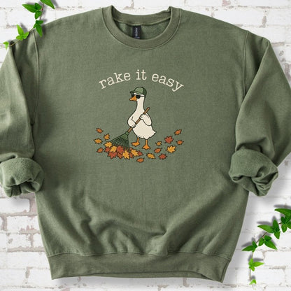 Military Green sweatshirt flat lay with ivory "rake it easy" lettering above a white goose in a green cap and sunglasses raking a pile of orange and gold fall leaves, on white brick with ivy sprigs.