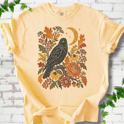 Butter T-shirt flat lay with a black raven, crescent moon and warm autumn flowers and foliage.