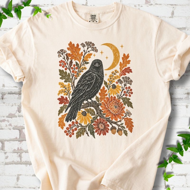 Ivory T-shirt flat lay with a black raven, crescent moon and warm autumn flowers and foliage.