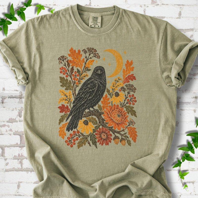 Khaki T-shirt flat lay with a black raven, crescent moon and warm autumn flowers and foliage.