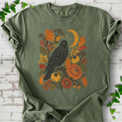 Moss T-shirt flat lay with a black raven, crescent moon and warm autumn flowers and foliage.