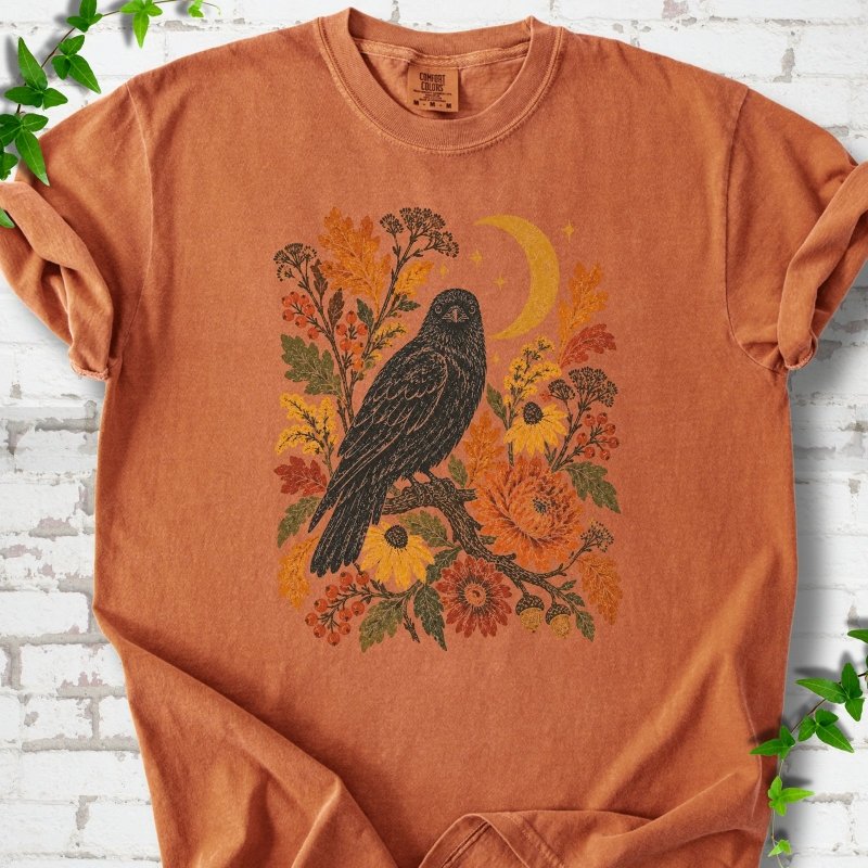 Yam T-shirt flat lay with a black raven, crescent moon and warm autumn flowers and foliage.