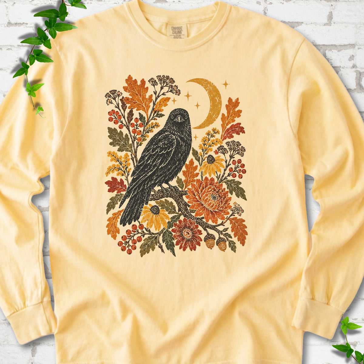 Butter long sleeve T-shirt flat lay with a black raven, crescent moon and warm autumn flowers and foliage.