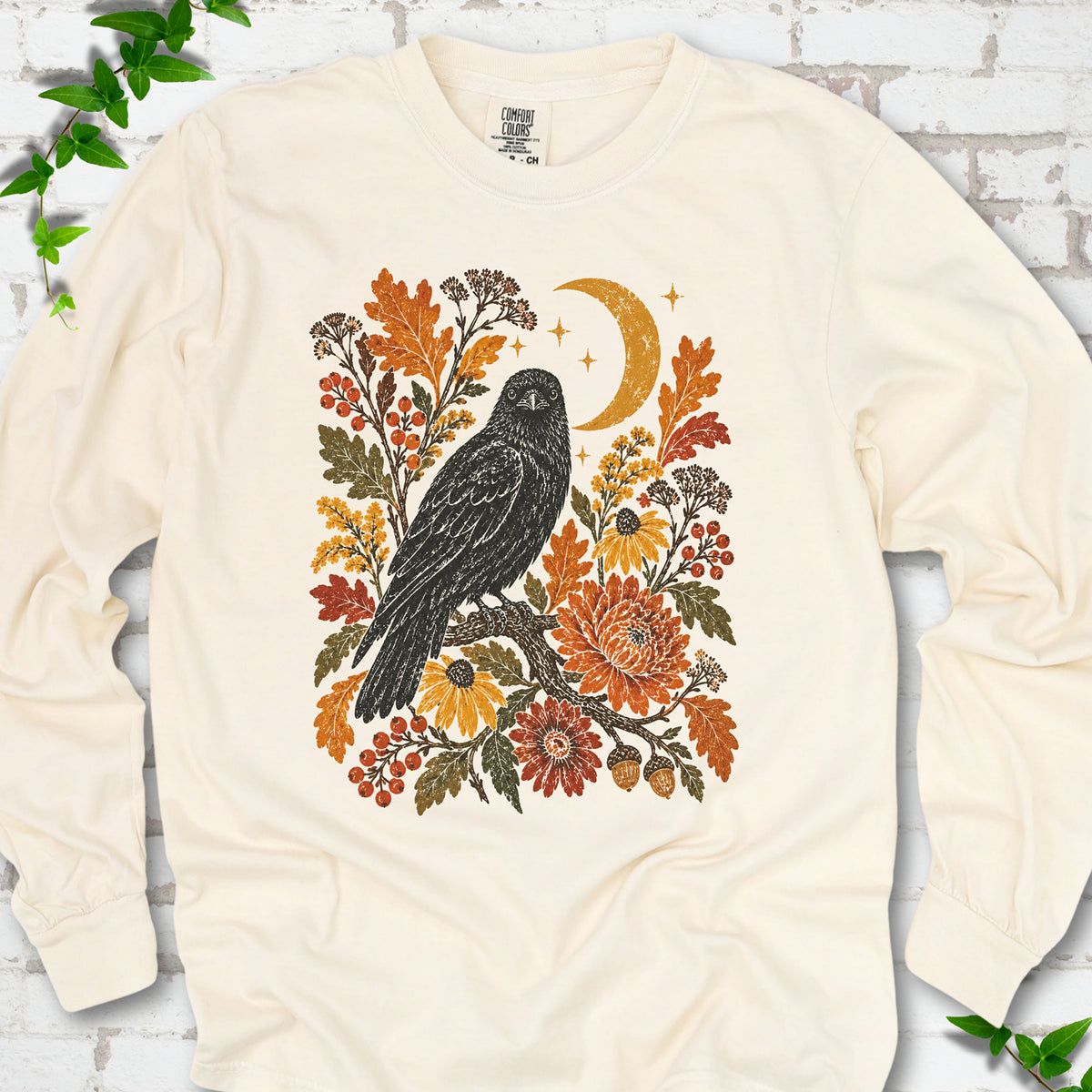 Ivory long sleeve T-shirt flat lay with a black raven, crescent moon and warm autumn flowers and foliage.