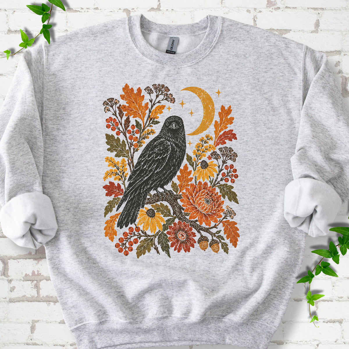 Ash crewneck sweatshirt flat lay with a black raven, crescent moon and warm autumn flowers and foliage.