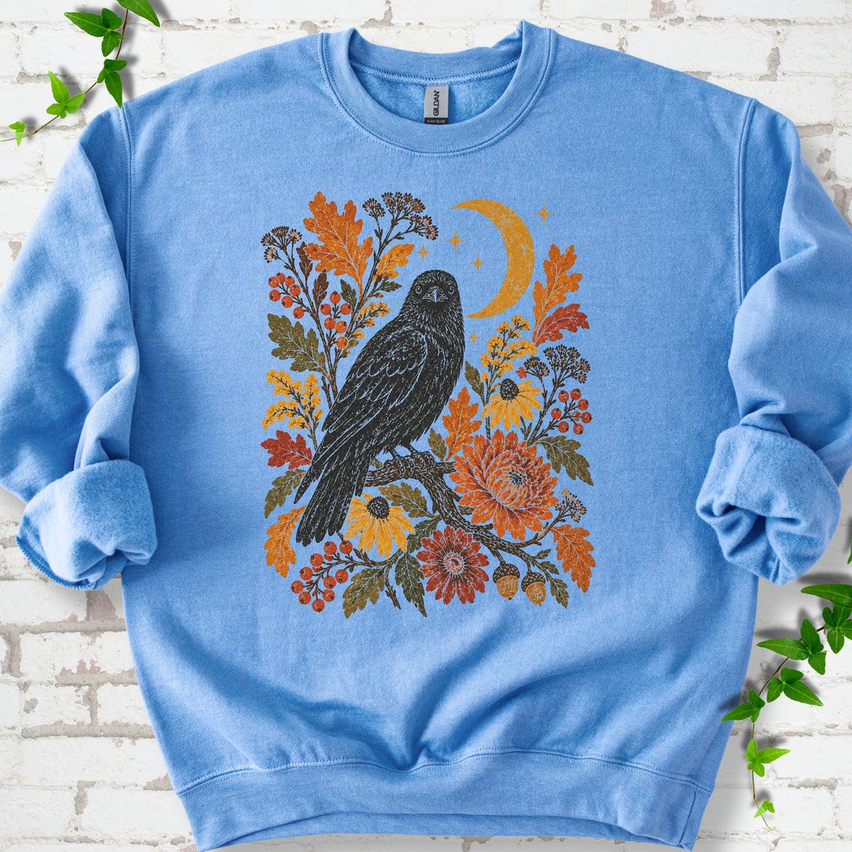 Carolina Blue crewneck sweatshirt flat lay with a black raven, crescent moon and warm autumn flowers and foliage.