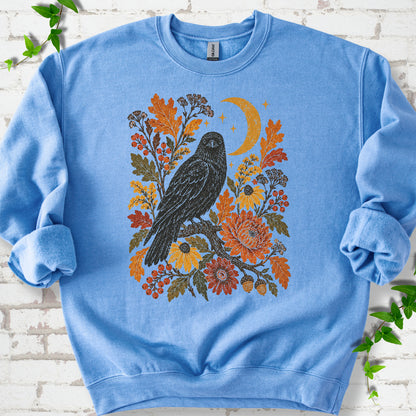 Carolina Blue crewneck sweatshirt flat lay with a black raven, crescent moon and warm autumn flowers and foliage.