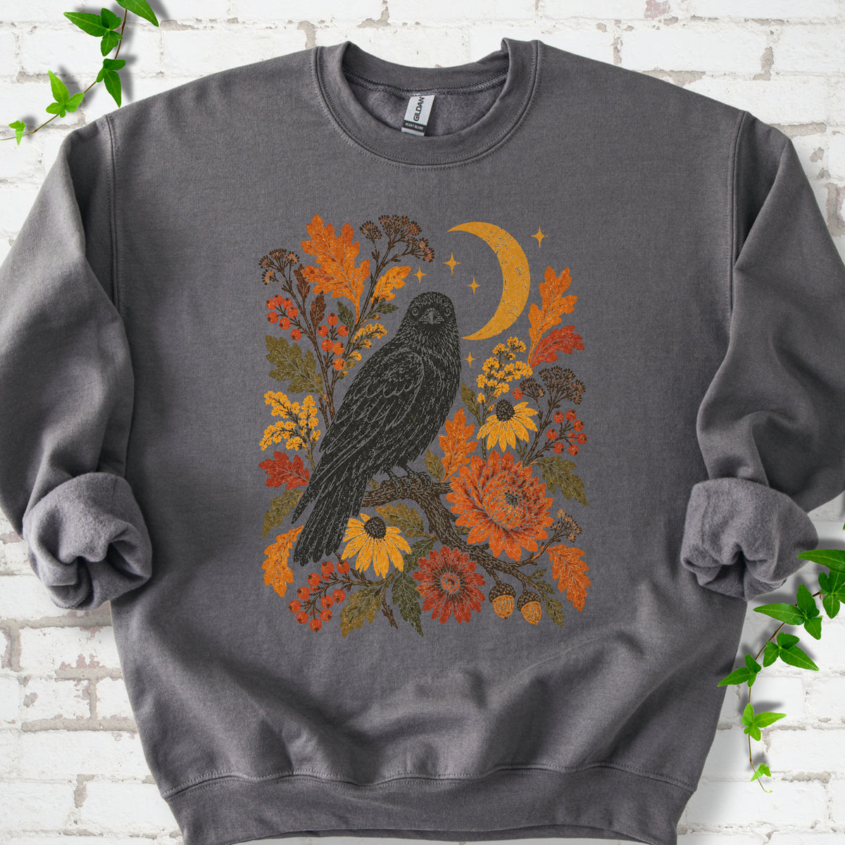 Charcoal crewneck sweatshirt flat lay with a black raven, crescent moon and warm autumn flowers and foliage.