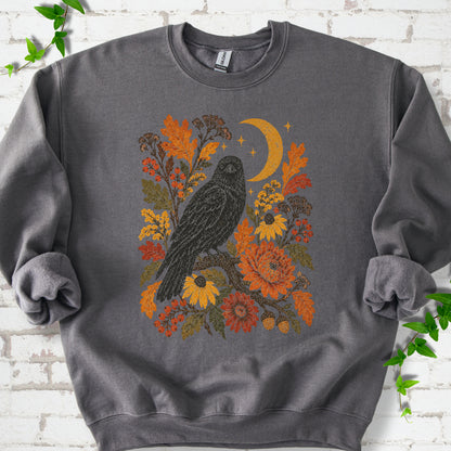 Charcoal crewneck sweatshirt flat lay with a black raven, crescent moon and warm autumn flowers and foliage.