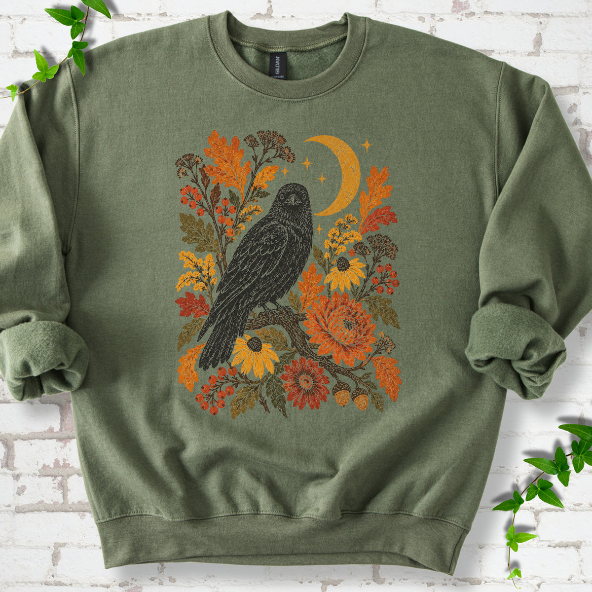 Military Green crewneck sweatshirt flat lay with a black raven, crescent moon and warm autumn flowers and foliage.