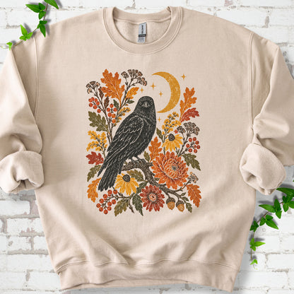 Sand crewneck sweatshirt flat lay with a black raven, crescent moon and warm autumn flowers and foliage.