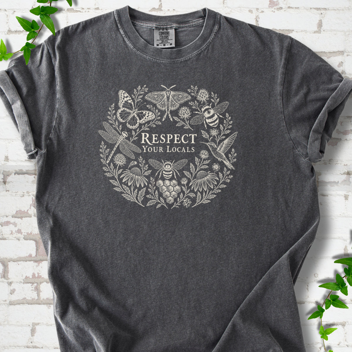 Pepper Comfort Colors® T-shirt flat lay featuring the words “Respect Your Locals” surrounded by pollinating insects, a hummingbird and flowers.