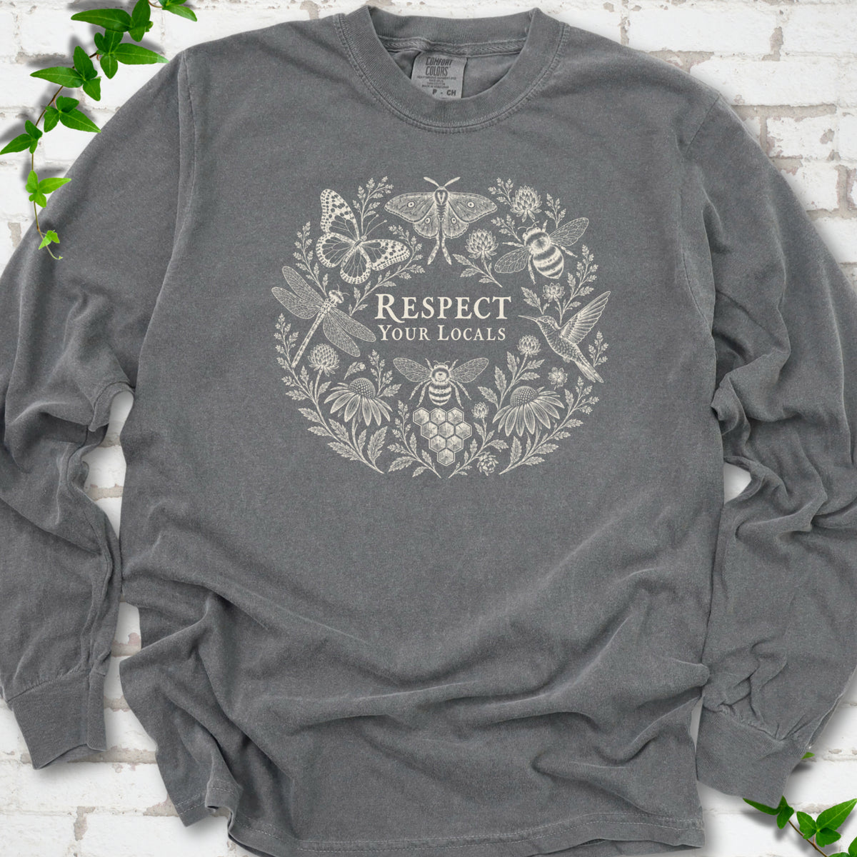 Pepper Comfort Colors® long sleeve tee flat lay featuring the words “Respect Your Locals” surrounded by pollinating insects, a hummingbird and flowers.