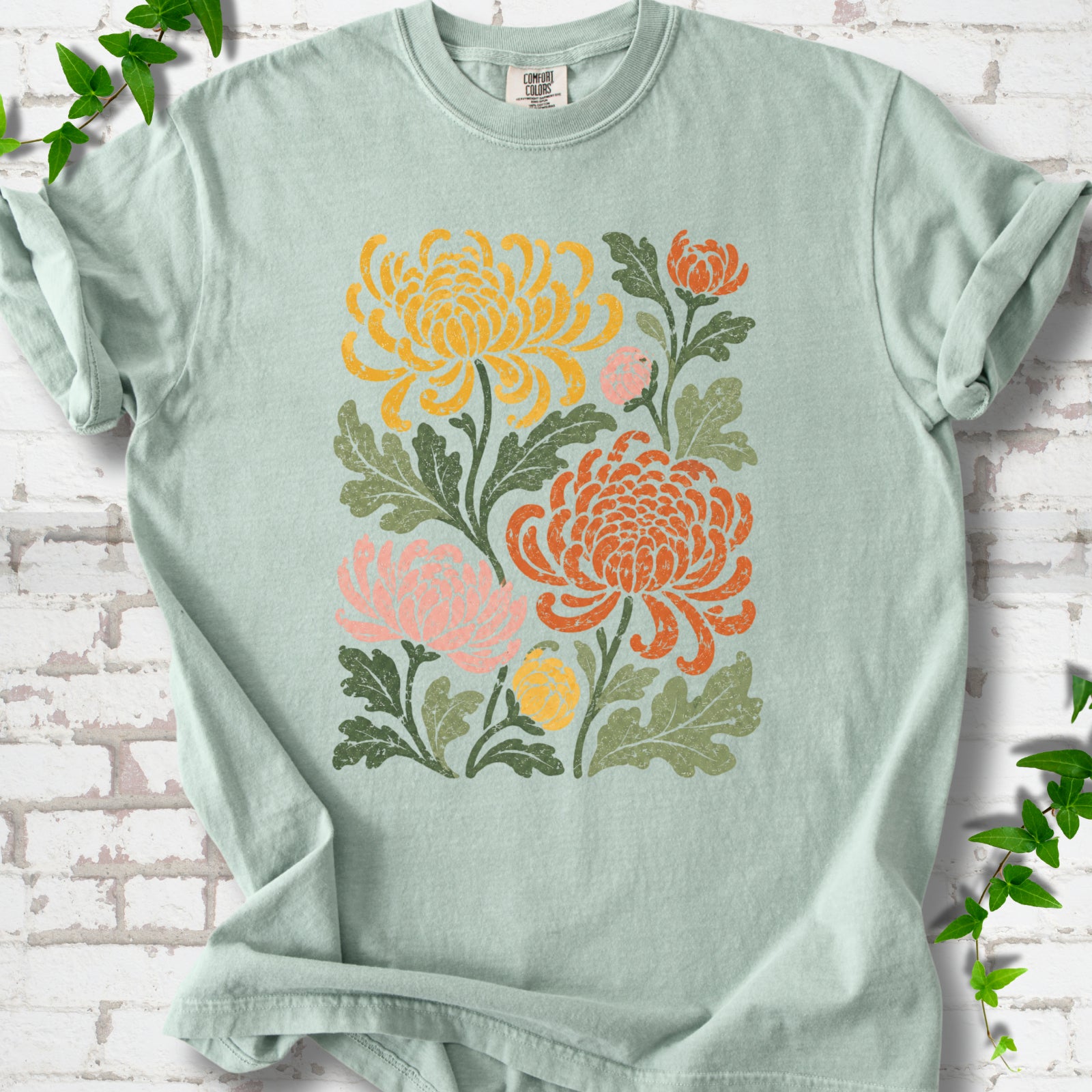 Bay Comfort Colors® short sleeve T-shirt flat lay with a distressed multicolor chrysanthemum garden graphic on a white brick background with ivy accents