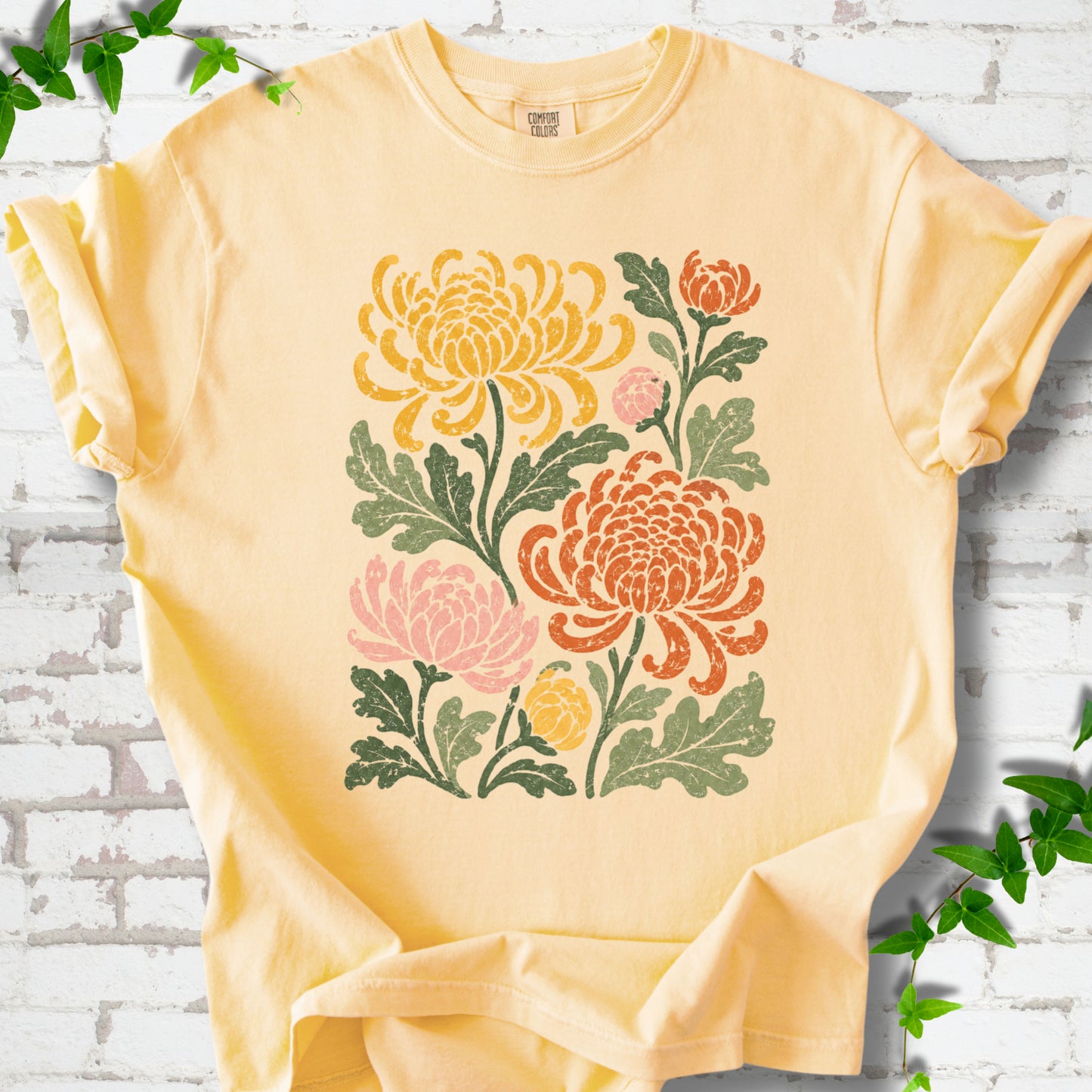 Butter Comfort Colors® short sleeve T-shirt flat lay with a distressed multicolor chrysanthemum garden graphic on a white brick background with ivy accents