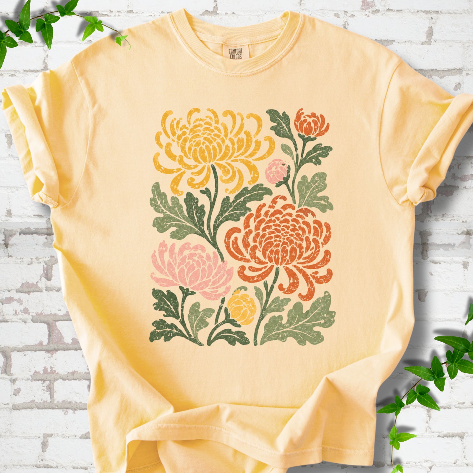Butter Comfort Colors® short sleeve T-shirt flat lay with a distressed multicolor chrysanthemum garden graphic on a white brick background with ivy accents