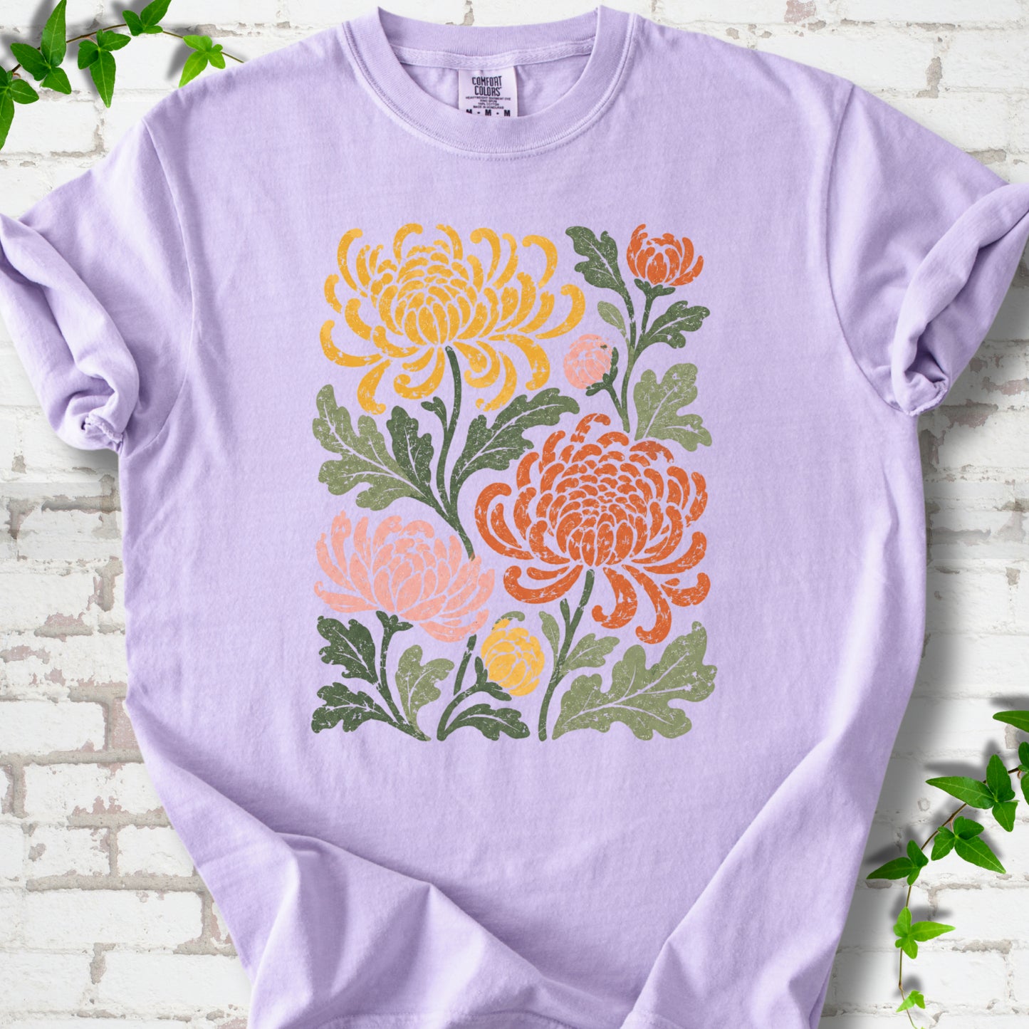 Orchid Comfort Colors® short sleeve T-shirt flat lay with a distressed multicolor chrysanthemum garden graphic on a white brick background with ivy accents