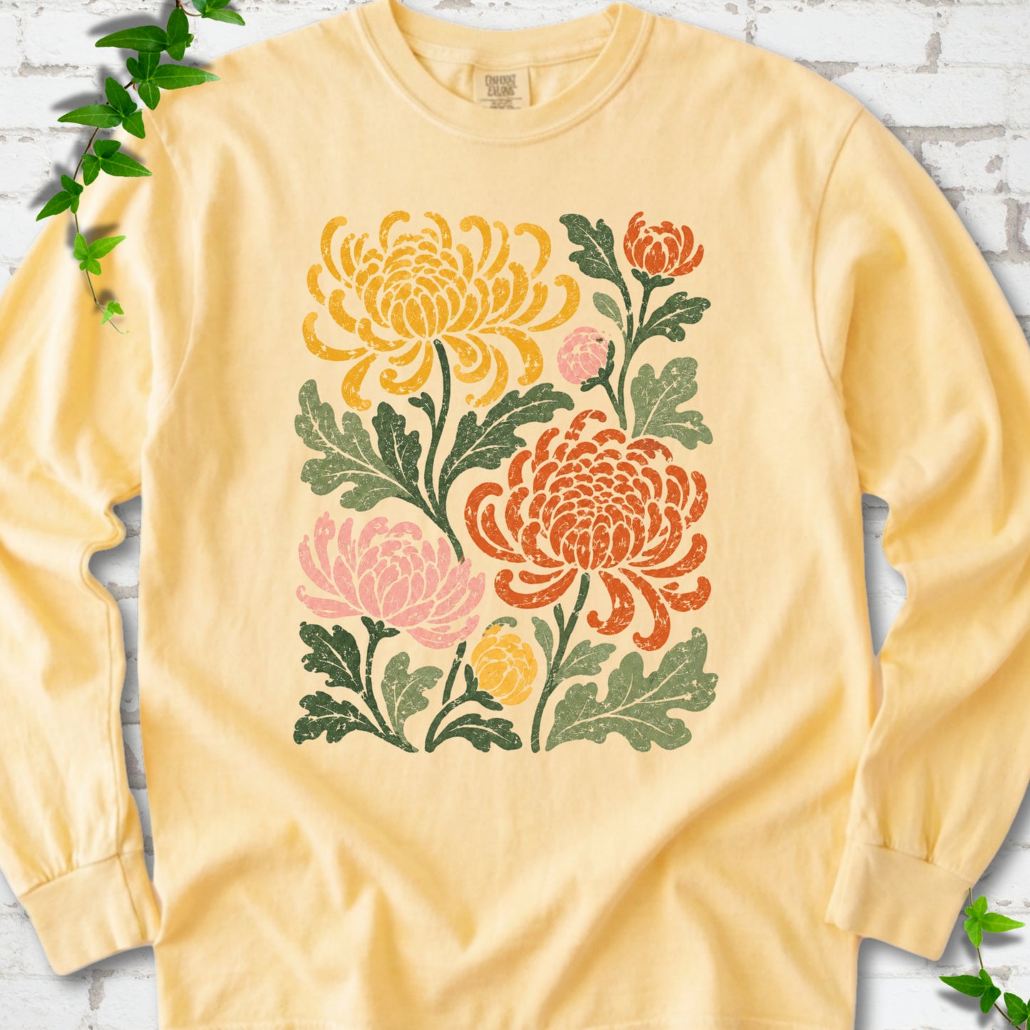 Butter Comfort Colors® long sleeve T-shirt flat lay with a distressed multicolor chrysanthemum garden graphic on a white brick background with ivy accents