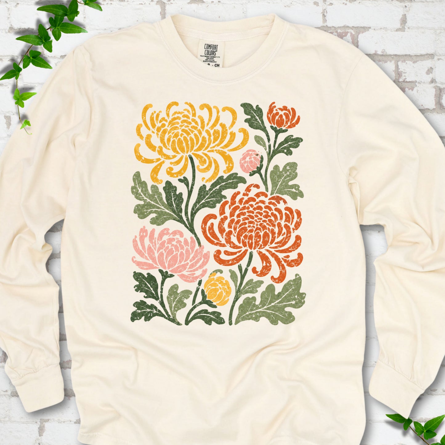 Ivory Comfort Colors® long sleeve T-shirt flat lay with a distressed multicolor chrysanthemum garden graphic on a white brick background with ivy accents