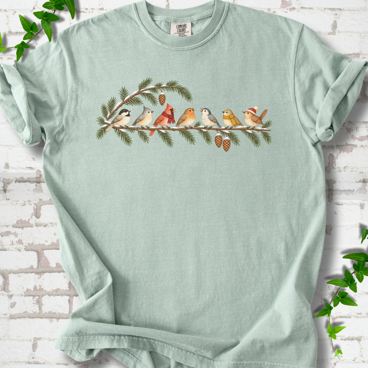 Bay T-shirt laid flat, featuring seven songbirds on snowy evergreen branches with pinecones. Some birds wear scarves, and one wears a knit hat.