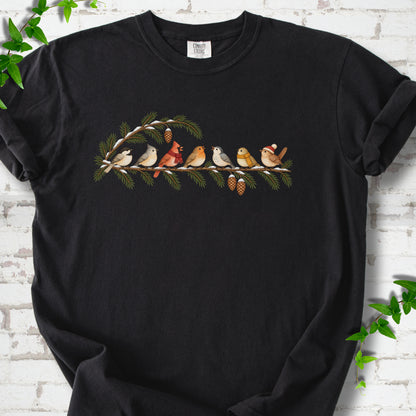 Black T-shirt laid flat, featuring seven songbirds on snowy evergreen branches with pinecones. Some birds wear scarves, and one wears a knit hat.
