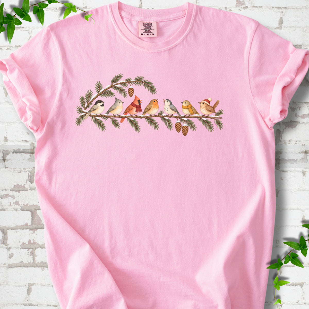 Blossom T-shirt laid flat, featuring seven songbirds on snowy evergreen branches with pinecones. Some birds wear scarves, and one wears a knit hat.