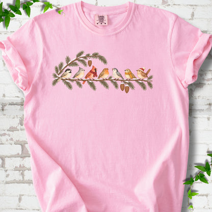 Blossom T-shirt laid flat, featuring seven songbirds on snowy evergreen branches with pinecones. Some birds wear scarves, and one wears a knit hat.