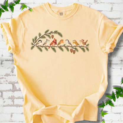 Butter T-shirt laid flat, featuring seven songbirds on snowy evergreen branches with pinecones. Some birds wear scarves, and one wears a knit hat.