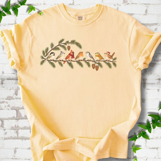 Butter T-shirt laid flat, featuring seven songbirds on snowy evergreen branches with pinecones. Some birds wear scarves, and one wears a knit hat.