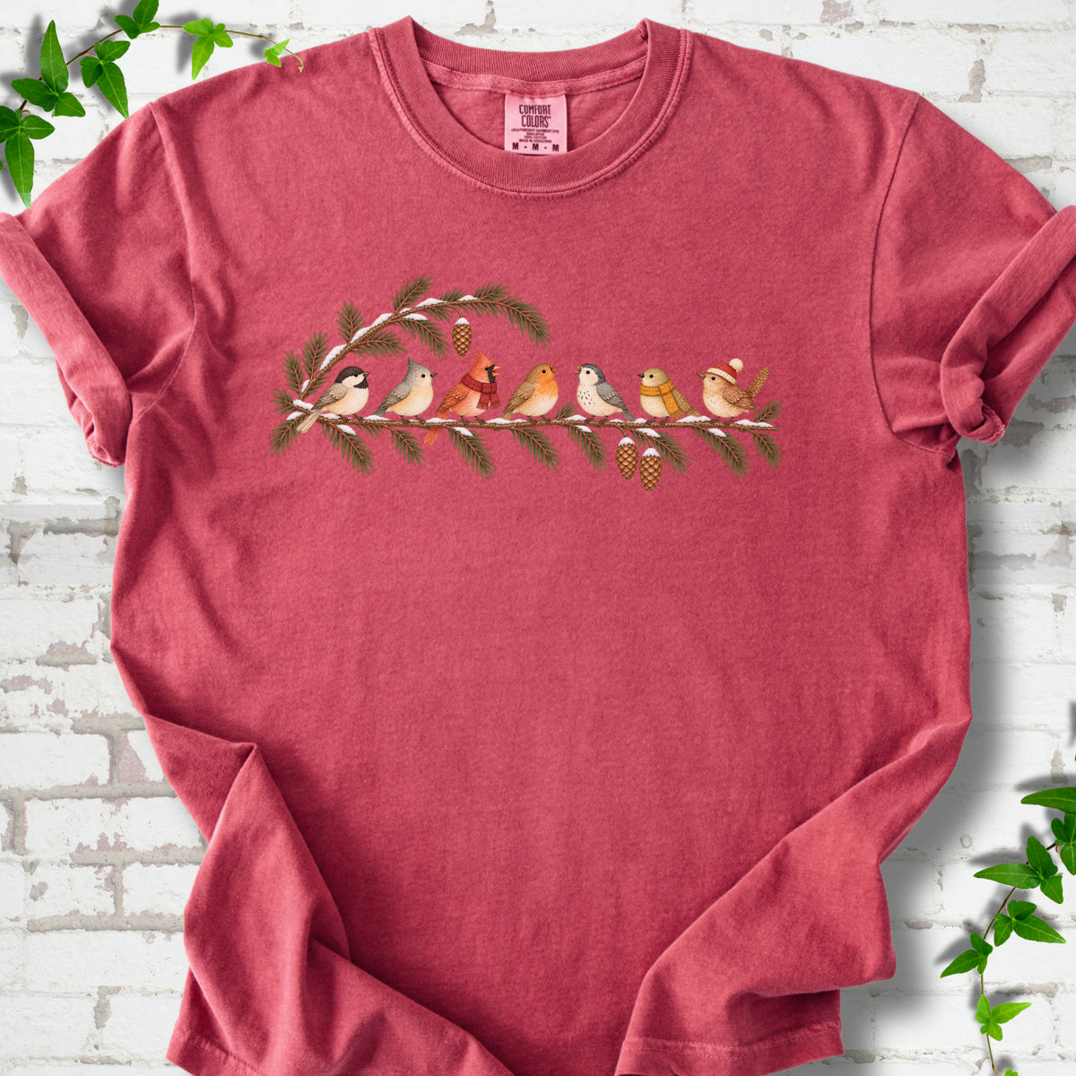 Crimson T-shirt laid flat, featuring seven songbirds on snowy evergreen branches with pinecones. Some birds wear scarves, and one wears a knit hat.