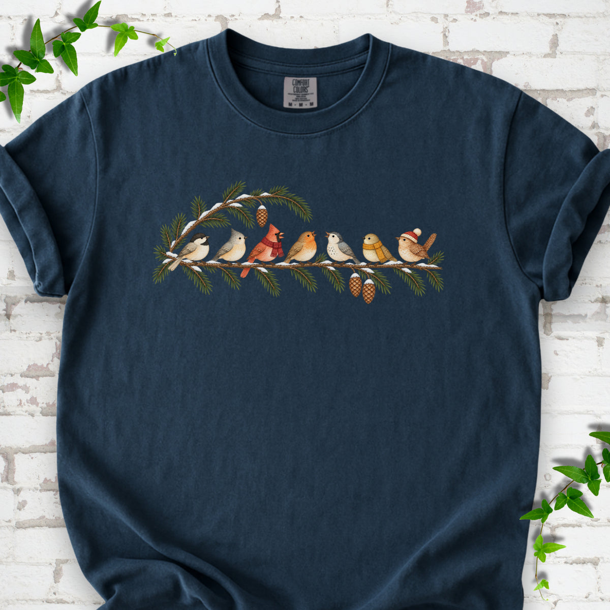 Midnight T-shirt laid flat, featuring seven songbirds on snowy evergreen branches with pinecones. Some birds wear scarves, and one wears a knit hat.
