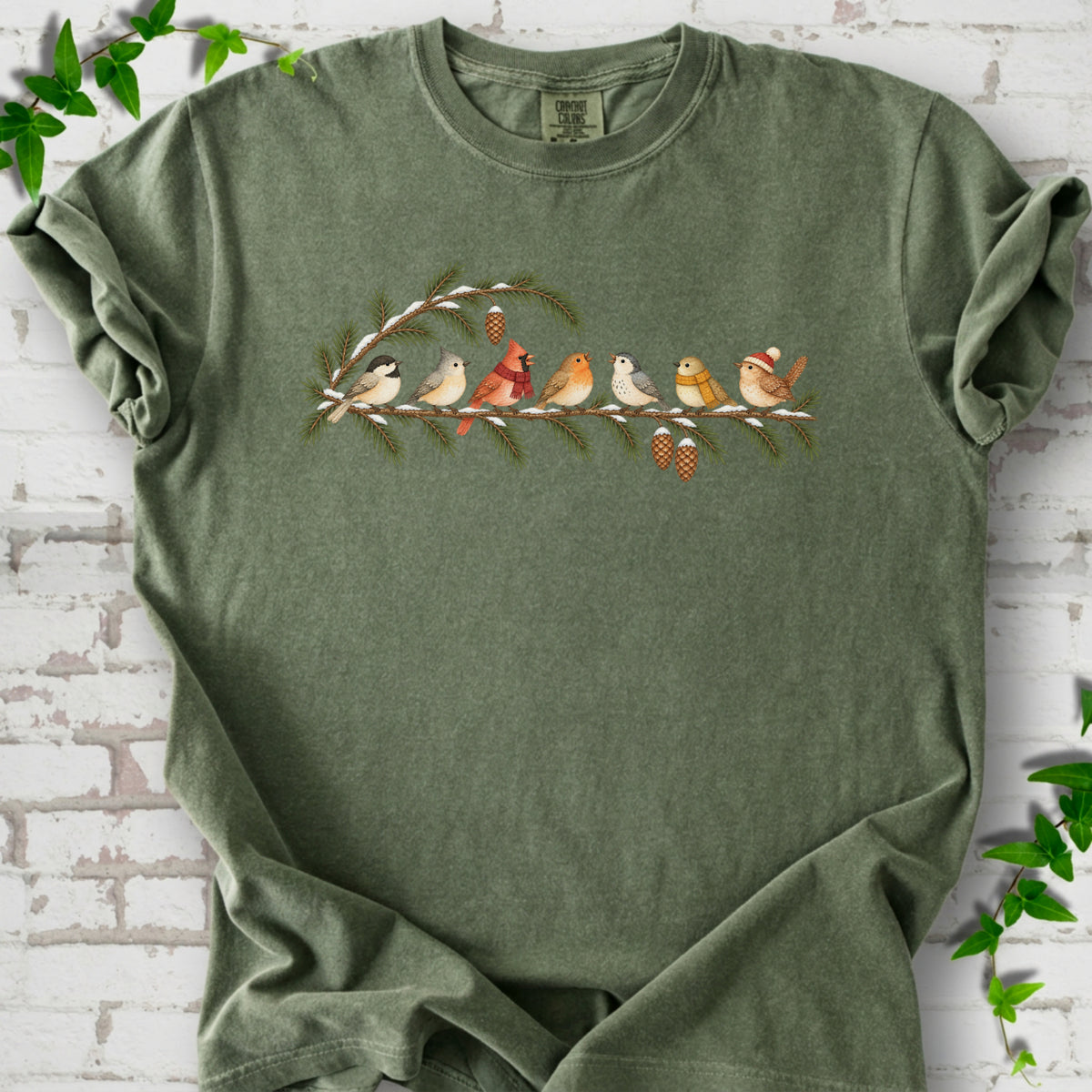Moss T-shirt laid flat, featuring seven songbirds on snowy evergreen branches with pinecones. Some birds wear scarves, and one wears a knit hat.