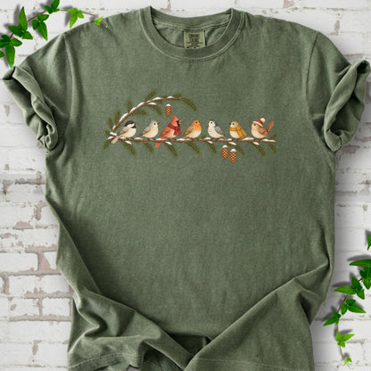 Moss T-shirt laid flat, featuring seven songbirds on snowy evergreen branches with pinecones. Some birds wear scarves, and one wears a knit hat.