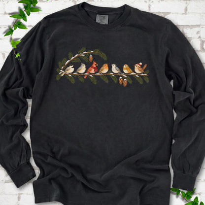Black long sleeve T-shirt laid flat, featuring seven songbirds on snowy evergreen branches with pinecones. Some birds wear scarves, and one wears a knit hat.