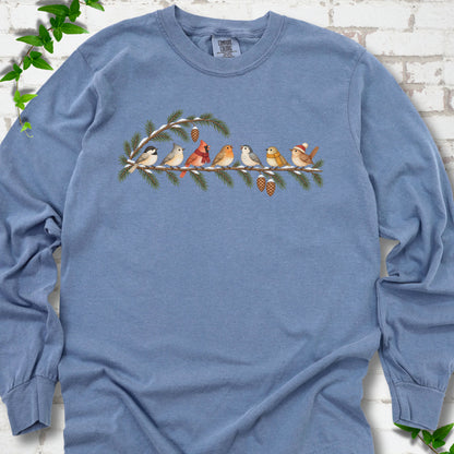 Blue Jean long sleeve T-shirt laid flat, featuring seven songbirds on snowy evergreen branches with pinecones. Some birds wear scarves, and one wears a knit hat.