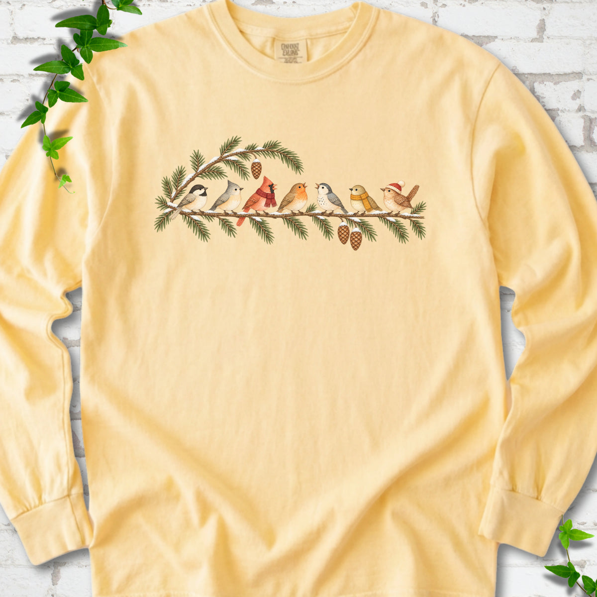 Butter long sleeve T-shirt laid flat, featuring seven songbirds on snowy evergreen branches with pinecones. Some birds wear scarves, and one wears a knit hat.