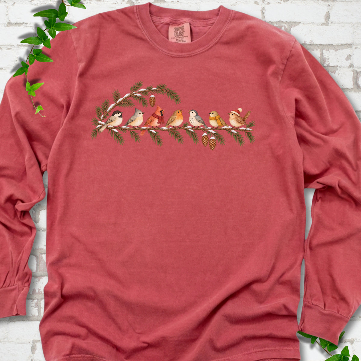 Crimson long sleeve T-shirt laid flat, featuring seven songbirds on snowy evergreen branches with pinecones. Some birds wear scarves, and one wears a knit hat.
