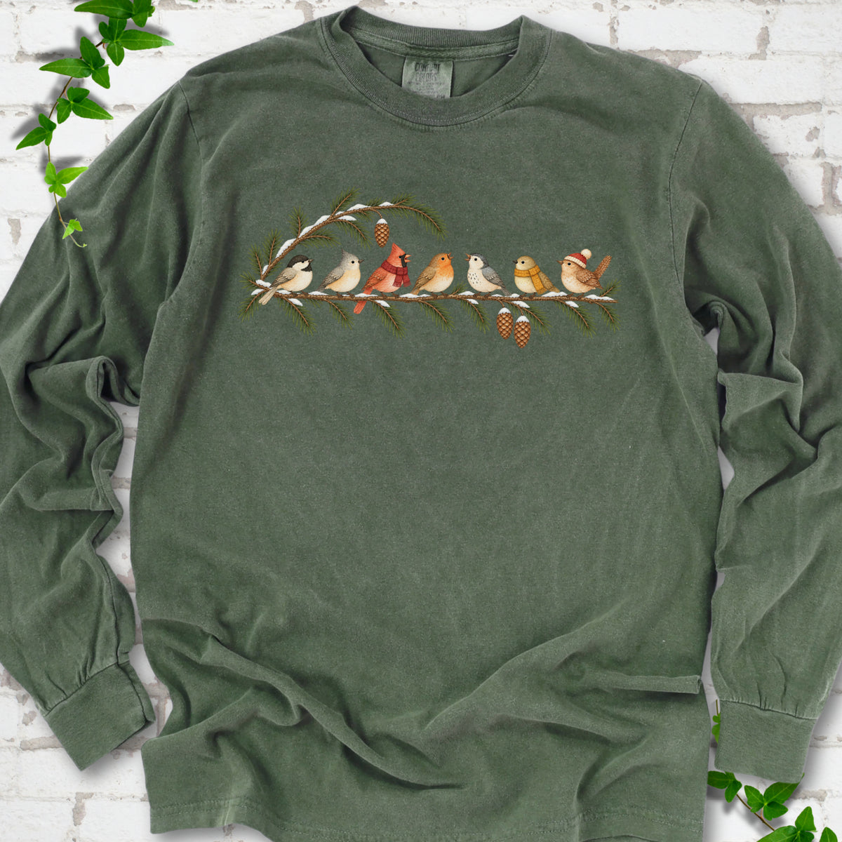 Hemp long sleeve T-shirt laid flat, featuring seven songbirds on snowy evergreen branches with pinecones. Some birds wear scarves, and one wears a knit hat.