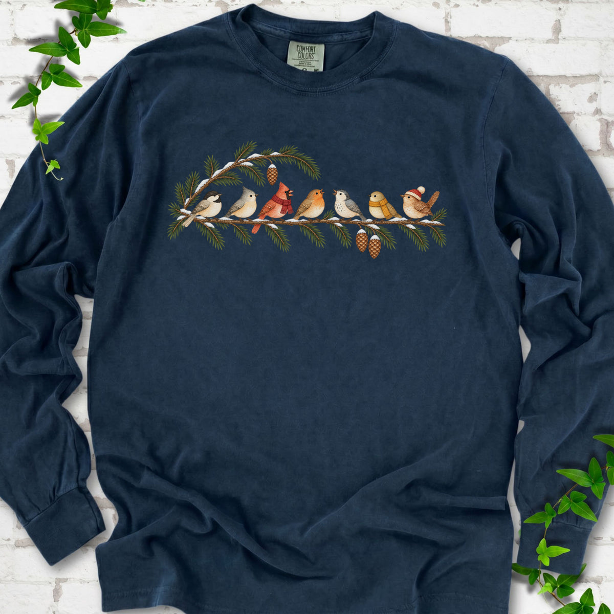 Midnight long sleeve T-shirt laid flat, featuring seven songbirds on snowy evergreen branches with pinecones. Some birds wear scarves, and one wears a knit hat.