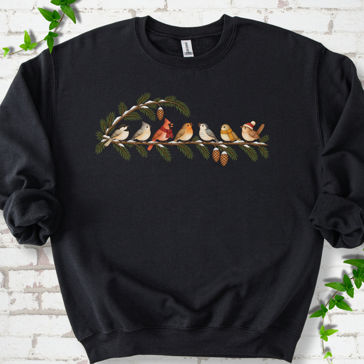 Black classic sweatshirt laid flat, featuring seven songbirds on snowy evergreen branches with pinecones. Some birds wear scarves, and one wears a knit hat.
