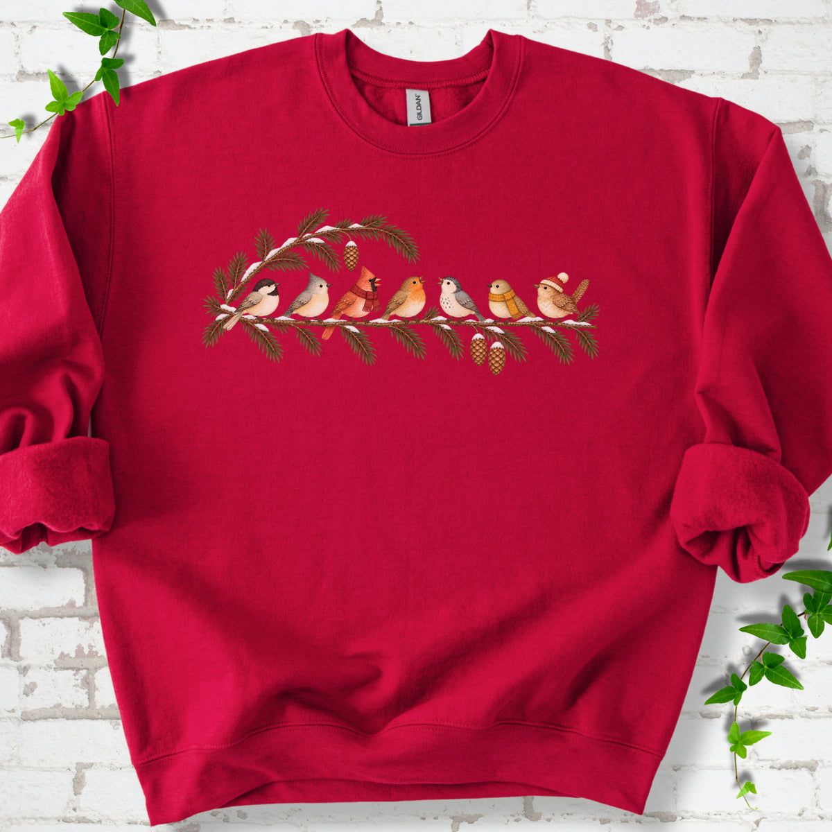 Cardinal Red classic sweatshirt laid flat, featuring seven songbirds on snowy evergreen branches with pinecones. Some birds wear scarves, and one wears a knit hat.