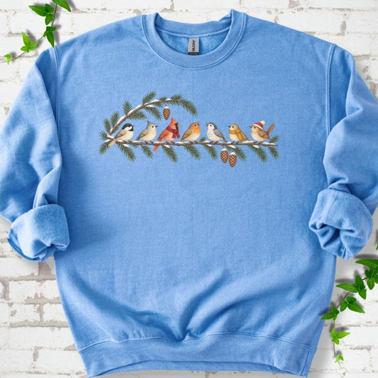 Carolina Blue classic sweatshirt laid flat, featuring seven songbirds on snowy evergreen branches with pinecones. Some birds wear scarves, and one wears a knit hat.