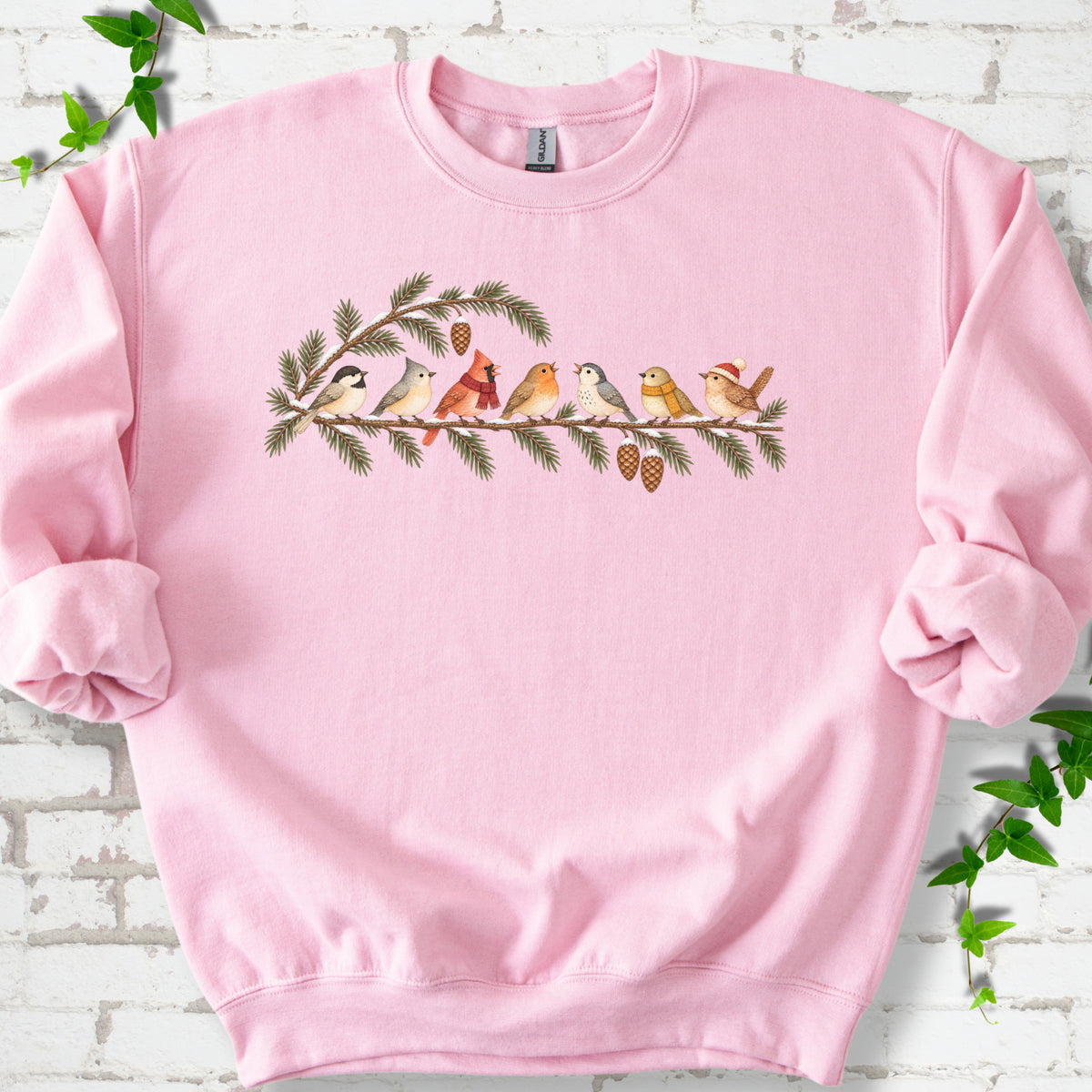 Light Pink classic sweatshirt laid flat, featuring seven songbirds on snowy evergreen branches with pinecones. Some birds wear scarves, and one wears a knit hat.