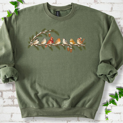 Military Green classic sweatshirt laid flat, featuring seven songbirds on snowy evergreen branches with pinecones. Some birds wear scarves, and one wears a knit hat.