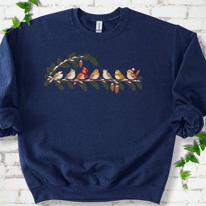 Navy classic sweatshirt laid flat, featuring seven songbirds on snowy evergreen branches with pinecones. Some birds wear scarves, and one wears a knit hat.