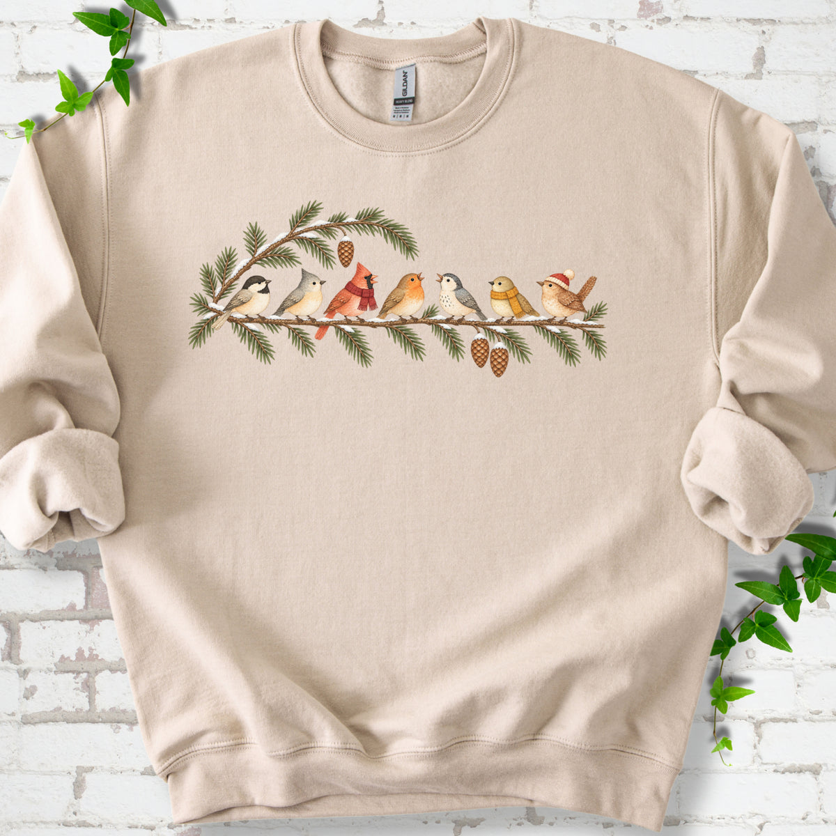 Sand classic sweatshirt laid flat, featuring seven songbirds on snowy evergreen branches with pinecones. Some birds wear scarves, and one wears a knit hat.