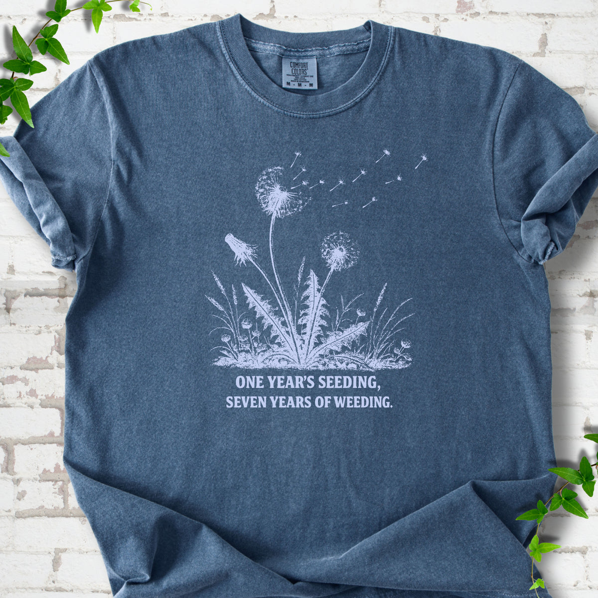 Denim T-shirt flat lay with dandelion seed heads, drifting seeds and 'One year's seeding, seven years of weeding' lettering.