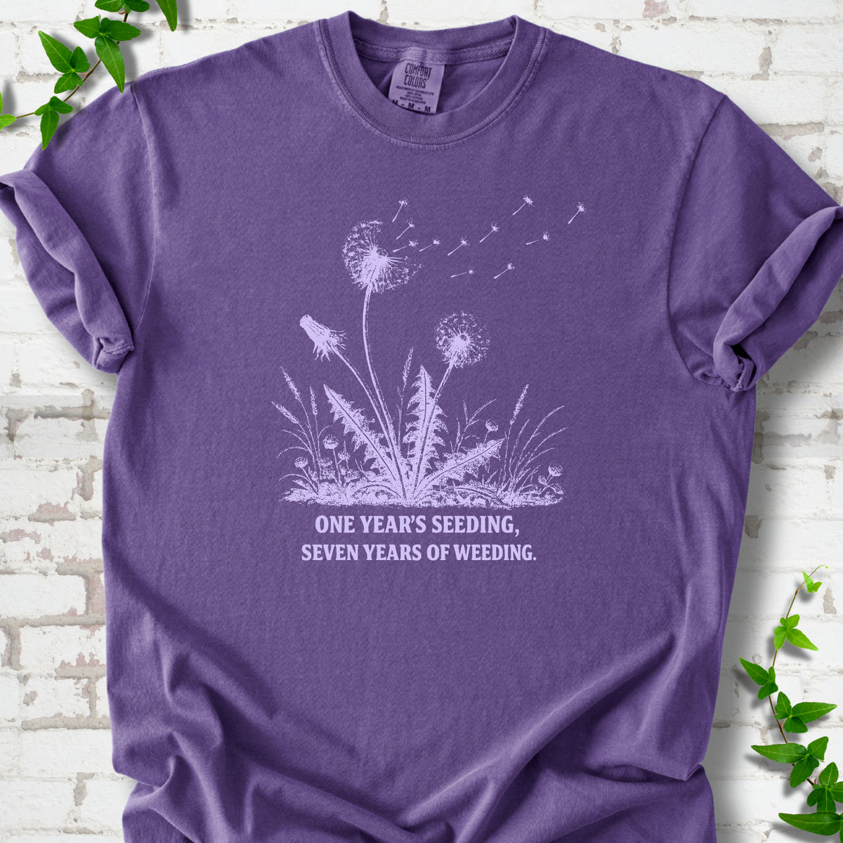 Grape T-shirt flat lay with dandelion seed heads, drifting seeds and 'One year's seeding, seven years of weeding' lettering.