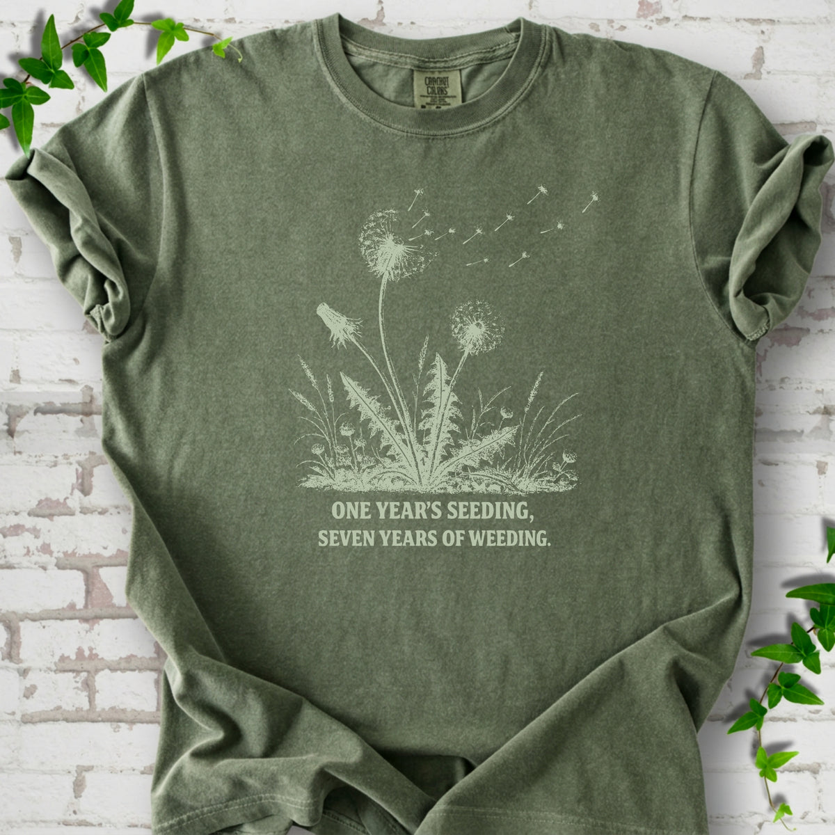 Moss T-shirt flat lay with dandelion seed heads, drifting seeds and 'One year's seeding, seven years of weeding' lettering.