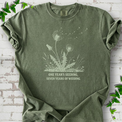 Moss T-shirt flat lay with dandelion seed heads, drifting seeds and 'One year's seeding, seven years of weeding' lettering.